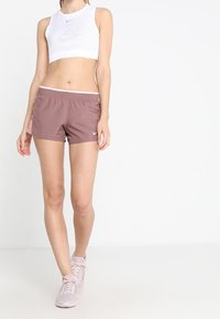 Weißes Netz-Crop-Top mit Logo, kombiniert mit pinkfarbenen Sportshorts. Schwarze Akzente an den Shorts, glatte Textur. Hellrosa Sportschuhe.