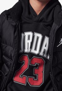 Giacca a piumino nera con colletto foderato in sherpa, indossata sopra una felpa nera con cappuccio con la scritta bianca "JORDAN" in grassetto e il numero "23" rosso.