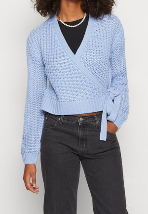 Femme portant un pull en maille bleu clair croisé noué à la taille sur un haut noir, associé à un jean noir taille haute et un collier de perles.