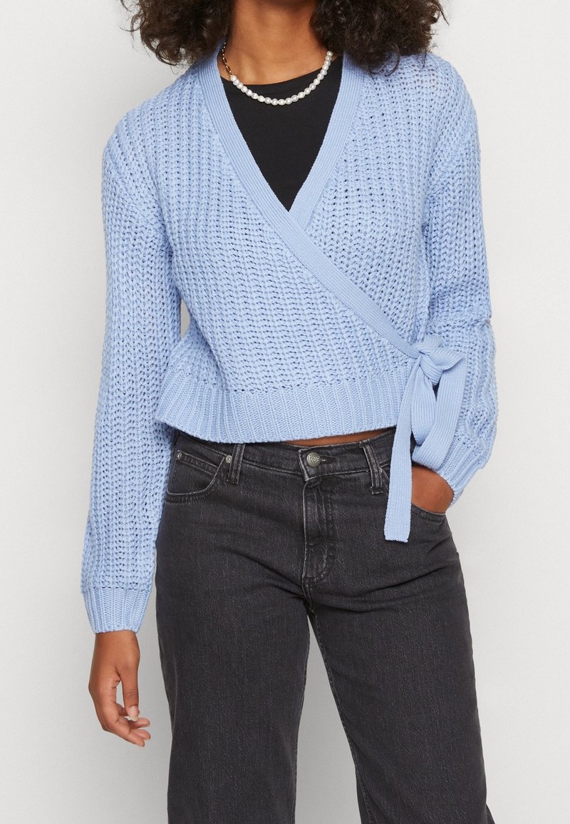 Femme portant un pull en maille bleu clair croisé noué à la taille sur un haut noir, associé à un jean noir taille haute et un collier de perles.