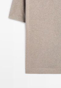 Beige strikket kortærmet sweater med ribbet kant ved ærmer og nederst på en hvid baggrund, hvor del af fronten og ærmet ses.