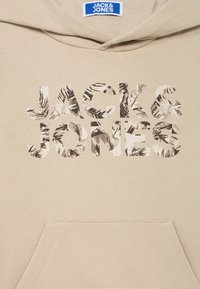 Sudadera con capucha beige con un gran logo recortado "JACK & JONES" con un patrón de hojas en su interior. Incluye un bolsillo delantero y detalle de capucha.