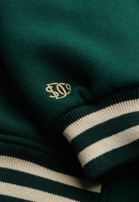Groene trui met crèmekleurige strepen op de manchetten, gemaakt van zacht materiaal, met een geborduurd logo in goud op de mouw.