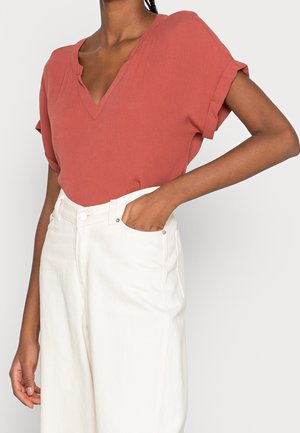 Vrouw draagt een roestkleurige korte mouw V-hals top, ingestopt in een witte broek met hoge taille, met één hand in de zak.