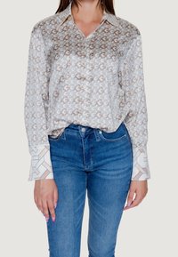 Blouse en soie avec un motif géométrique beige et blanc, dotée d'un col pointu et de larges poignets. Associée à un jean denim bleu.