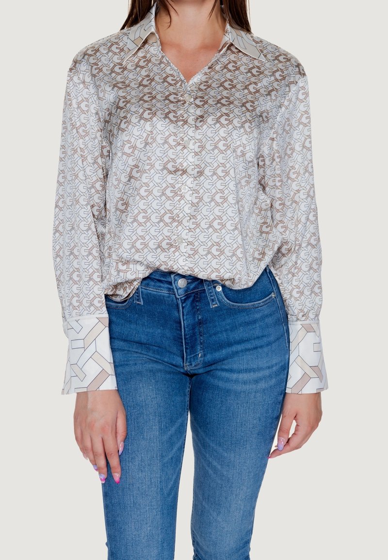 Blouse en soie avec un motif géométrique beige et blanc, dotée d'un col pointu et de larges poignets. Associée à un jean denim bleu.
