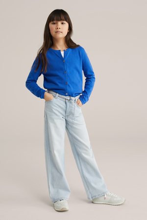 Niña de pie con las manos en los bolsillos de unos jeans azul claro, vistiendo un cárdigan abotonado azul brillante y zapatillas blancas sobre un fondo neutro.