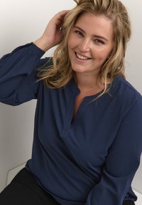 Námořnická modrá, dlouhá blůza s dlouhým rukávem a výstřihem do V s detailem knoflíků. Měkká, strukturovaná tkanina s volným střihem, kombinována s černými kalhotami.