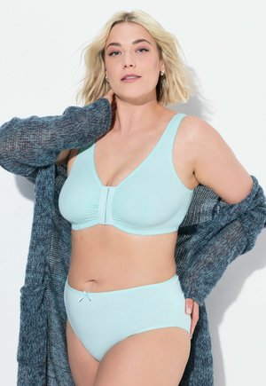 Mintgroene beha met een V-hals ontwerp en een geplooid voorpaneel, gecombineerd met bijpassende hoge tailleslips. Getextureerde blauwe cardigan overheen gedrapeerd.