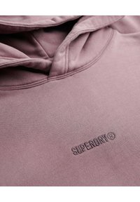 Sudadera ligera de color púrpura hecha de tela suave, con un logo "SUPERDRY" bordado y capucha con cordones. Textura suave y diseño casual.