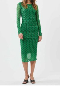 Robe verte à manches longues avec un motif de cactus, silhouette ajustée et longueur midi. Tissu extensible à texture transparente. Sandales noires.