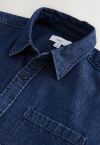 Chemise en denim bleu foncé avec col pointu, deux boutons noirs et poche poitrine, étalée à plat sur une surface blanche.