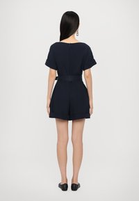 Marineblå jumpsuit med korte ermer, belte i midjen og oppbrettede mansjetter. Har et nøkkelhull bak og jevn, lettvektig stoff.