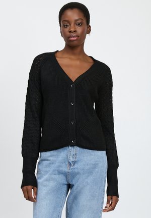 Zwarte gebreide cardigan met een V-hals, voorzien van textuurmouwen en zes knoopsluitingen. Gepaarde met lichtblauwe denimbroeken.