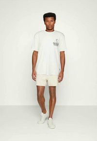 Homem de pé em calções bege, T-shirt gráfica branca com uma pequena impressão do Snoopy e ténis brancos em frente a um fundo claro e liso.