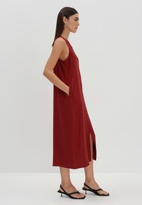 Rotes ärmelloses Kleid mit seitlichen Taschen, Seitenschlitz und lässiger Passform; aus leichtem Stoff gefertigt, ergänzt durch schwarze Slipper.