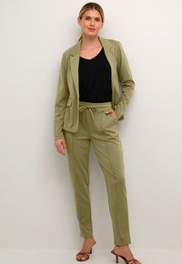 Svetlo zeleni suknjič s prilagojenim krojem, over črnim vrhom z V-izrezom, v kombinaciji s usklajenimi hlačami v slogu joga in rjavimi sandalami s peto.