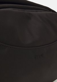 Sac en tissu noir avec une texture lisse, arborant un logo de marque discret. Le design est minimaliste avec des lignes épurées et un simple accent de poche.