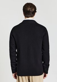 Schwarze Strickjacke mit Schalkragen, verlängerten Ärmeln und geradem Saum. Texturiertes Material mit feinen Stickdetails.