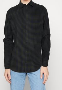 Camicia button-up a quadri verde scuro e blu navy con maniche lunghe, colletto e bottoni neri. Indossata con jeans azzurri chiari.