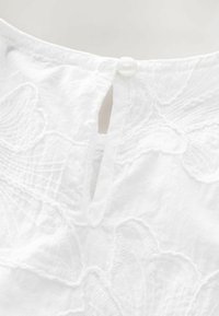 Top bianco in cotone con ricami floreali, apertura a chiave sul retro e bottone bianco rotondo per la chiusura. Textura liscia.