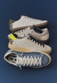 Quatre paires de baskets basses en blanc, gris et nuances de bleu disposées en diagonale sur un fond bleu foncé.