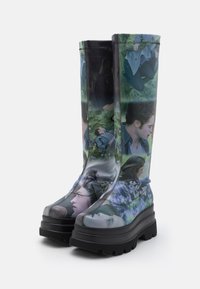 Vyšší gumové holínky vyrobené z lesklého materiálu, které mají koláž tváří a přírody, s tlustou černou gumovou podrážkou a texturovaným gripem.