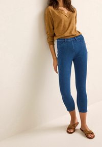 Haut marron à manches longues avec un col en V, associé à un jean bleu moulant longueur cheville et des sandales plates marron avec un design de lanière torsadée.