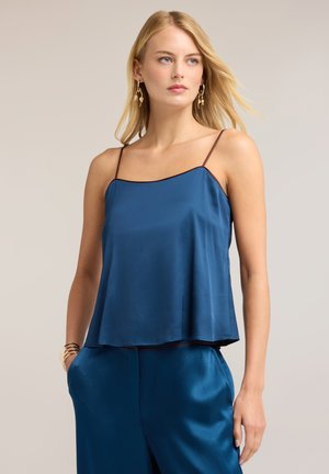 Jonge vrouw met blond haar die een blauwe satijnen spaghetti-strap top en bijpassende broek draagt, geaccentueerd met gouden oorbellen en armbanden.