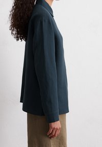 Camicia verde scuro a maniche lunghe in un tessuto leggero, con colletto, polsini abbottonati e vestibilità rilassata, indossata con una gonna marrone chiaro.