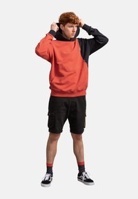 Felpa a blocchi di colore rossa e nera con finitura testurizzata, abbinata a pantaloni corti e sneakers, dotata di cappuccio con cordoncino e dettagli di cucitura.