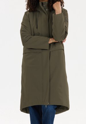 Persoon die een olijfgroene lange winterjas met capuchon draagt en een blauwe jeans, met gekruiste armen en een hand die de kin raakt.