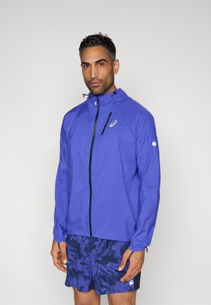 Lekka kurtka w kolorze królewskiego niebieskiego z zamkiem z przodu, regulowanym kapturem i czarnymi akcentami, zestawiona z niebieskimi wzorzystymi szortami.