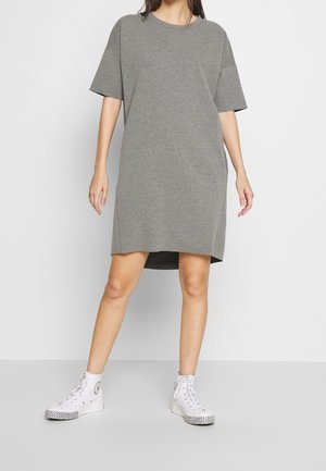 Kvinde iført en løs grå kortærmet kjole og hvide høje sneakers står mod en ensfarvet hvid baggrund.