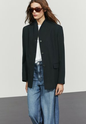 Frau trägt einen dunkel oversized Blazer, weißes Top, hellblaue lockere Jeans und große braune Sonnenbrille und steht vor einfachem Hintergrund.