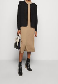 Cardigan nero peloso, vestito beige lungo fino al ginocchio, stivali neri alla caviglia con tacco alto e una borsa strutturata bianco e nero.