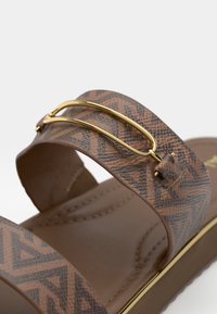 Bride de sandale marron avec motif géométrique et détail barre en métal doré sur une semelle intérieure douce et matelassée, avec un détail assorti sur le bord de la semelle.