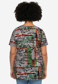 Grafische t-shirt met een mix van kleuren—groen, oranje en roze—op een grijze achtergrond, met abstracte patronen en handgeschreven tekst. Korte mouwen.