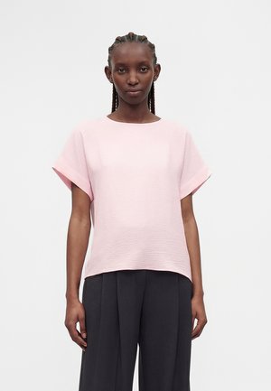 Jong persoon met gevlochten haar, een lichtroze top met korte mouwen en losse zwarte broek dragend, staand tegen een effen achtergrond.