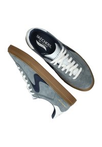 Zapatillas de ante grises con cordones blancos y detalles en azul marino, que presentan una suela de goma texturizada y la marca "SKECHERS" en la lengüeta y el talón.