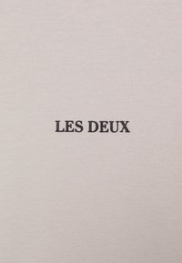 Weißes Baumwoll-T-Shirt mit aufgesticktem Text "LES DEUX" in Marineblau. Glatte Textur mit einem einfachen, minimalistischen Design.