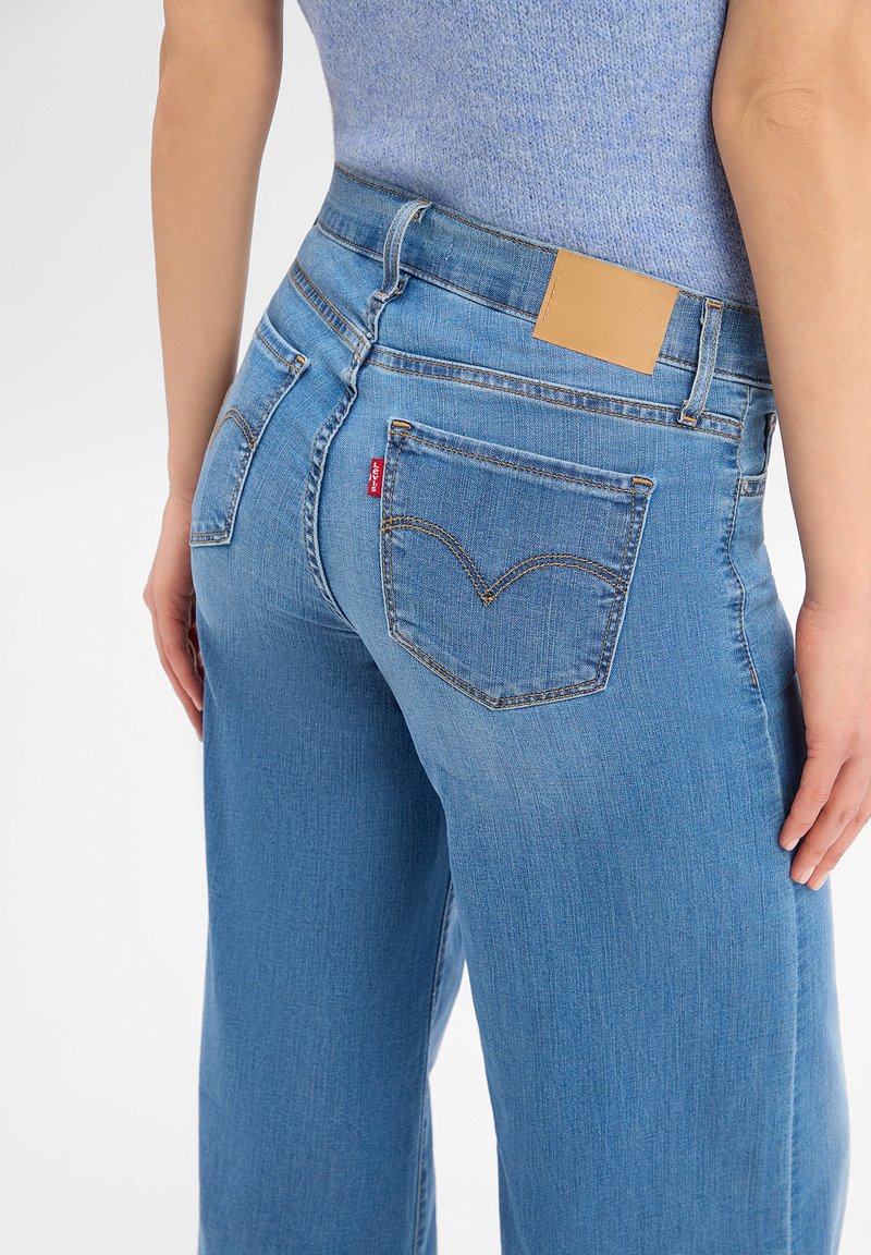 Woman wearing light blue denim jeans and a sleeveless knit top, with hands relaxed by her sides, shown from the back waist down.