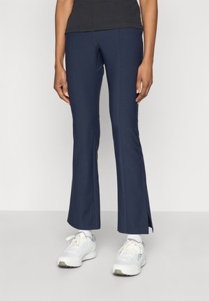 Pantalon évasé bleu marine avec une texture lisse, doté d'ouvertures latérales à l'ourlet. Association avec un haut noir et des chaussures de sport blanches.