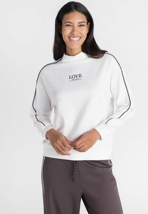 Lachende vrouw met lang donker haar die een witte trui met zwarte strepen en tekst "LOVE IS MY STYLE," draagt en donkere broek met witte strepen.