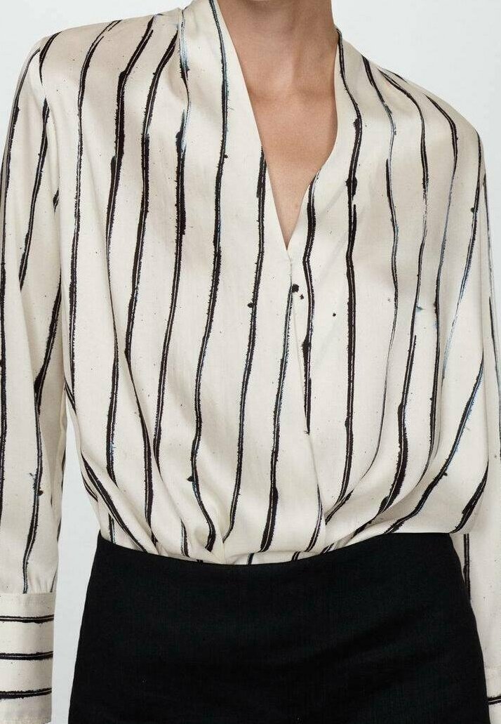Blouse en soie avec un col en V profond, ornée de rayures verticales noires sur un fond crème. Coupe fluide et drapée, détail de boutons sur le devant.