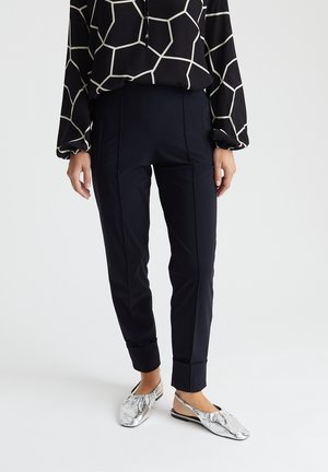 Person wearing black tailored pants with cuffed hems, a black and white geometric patterned blouse, and silver flat shoes with slingback straps.