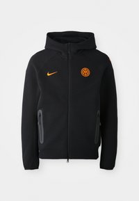 Sudadera negra con cremallera frontal, logo de Nike naranja y emblema circular naranja. Cuenta con dos bolsillos laterales con cremallera y un diseño texturizado.