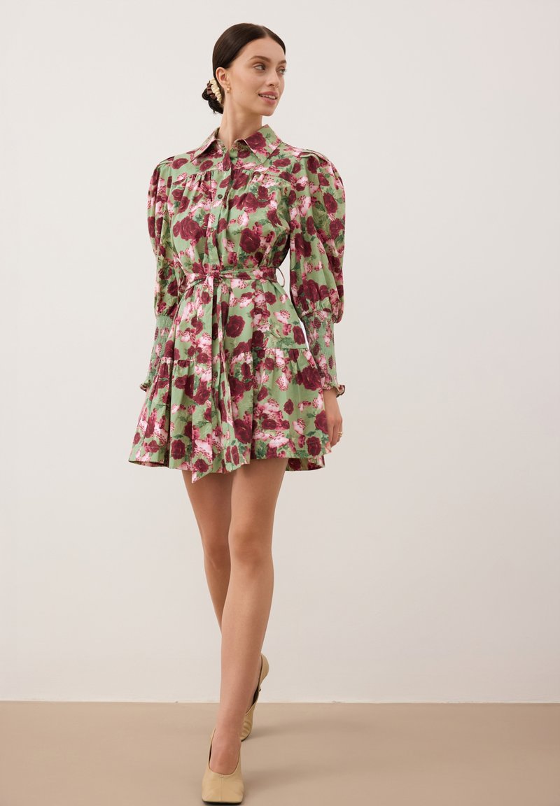 Kwiatowa sukienka w odcieniach zieleni z różowymi i burgundowymi wzorami, z bufiastymi rękawami, zapinana na guziki z przodu oraz z wcięciem w talii podkreślonym paskiem.
