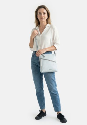 Frau trägt weiße Bluse und blaue Jeans, hält eine hellgraue Umhängetasche mit Reißverschluss und steht vor weißem Hintergrund.