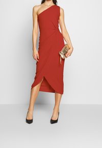 Röd axelklänning med knutdetalj vid midjan, med en hög slit vid låret. Kombinationen med en gyllene kuvertväska och svarta klackar.